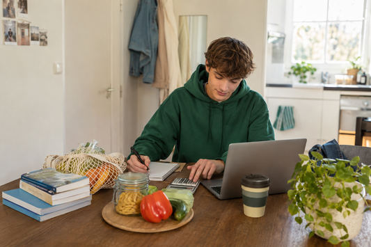 Student maakt thuis een budget met een laptop, rekenmachine, studieboeken en boodschappen.